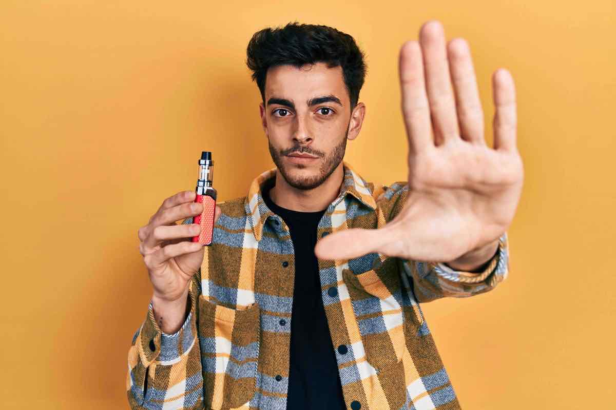Young hispanic man holding electronic cigarette with open hand doing stop